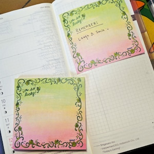 May include: An open planner with a grid page and a weekly calendar page. Two square sticky notes with a green-to-pink gradient, a dark vine border with clover accents, and the text "i'm so lucky!" are placed on the pages. One note reads "REMEMBER: Laugh & Smile".