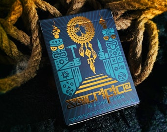 Puede incluir: Una baraja de cartas con un diseño azul y dorado. El dorso de la carta presenta un diseño tribal con una máscara tiki y un atrapasueños. El texto "Sacrifice Playing Cards" está impreso en dorado en la parte inferior de la carta.