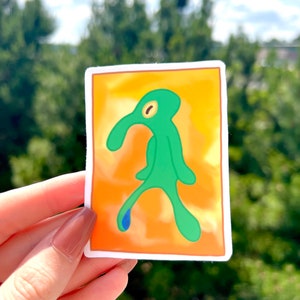 May include: A sticker featuring a green, cartoon character with a long nose and a single eye. The character is standing on an orange background with a blue teardrop.