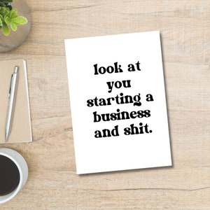 以下が含まれることがあります： 「look at you starting a business and shit」と書かれた黒い文字の白いカード。カードは、ペン、メモ帳、小さな植物、コーヒーカップのある木製の机の上にあります。