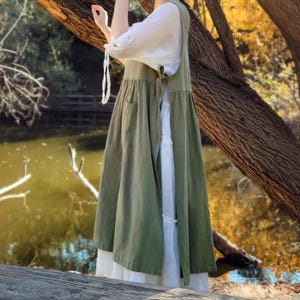 May include: An olive green apron dress with pockets, layered over a white dress. The dress has a relaxed fit and is tied at the back. The sleeves of the white dress are long and gathered at the wrists. The background features a lake and trees.