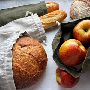 May include: An overhead shot features fresh bread and apples in reusable produce bags. Two baguettes and a round loaf of bread are visible, along with three red and yellow apples. The bags are in shades of green and beige, suggesting an eco-friendly theme.