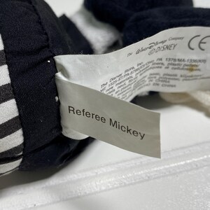 May include: Close-up of a Referee Mickey plush toy. The toy is black and white, with a tag that reads "Referee Mickey". The tag also includes the Disney logo and other text.
