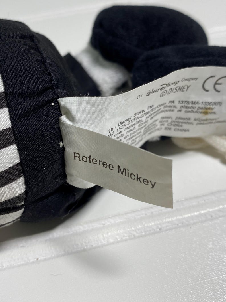 May include: Close-up of a Referee Mickey plush toy. The toy is black and white, with a tag that reads "Referee Mickey". The tag also includes the Disney logo and other text.