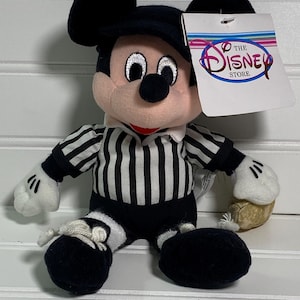 May include: A plush Mickey Mouse toy wearing a black and white striped referee shirt, black shorts, and a black hat. The toy has white gloves and shoes, and a tag that says "The Disney Store".