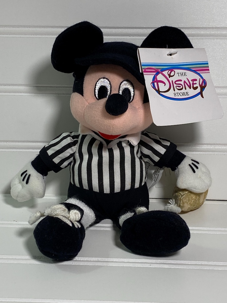May include: A plush Mickey Mouse toy wearing a black and white striped referee shirt, black shorts, and a black hat. The toy has white gloves and shoes, and a tag that says "The Disney Store".