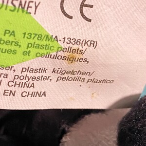 May include: Close-up of a white tag with the Disney logo and a CE mark. The tag includes text in multiple languages, including English, French, and Spanish, with the text "PA 1378/MA-1336(KR)" and "plastic pellets".