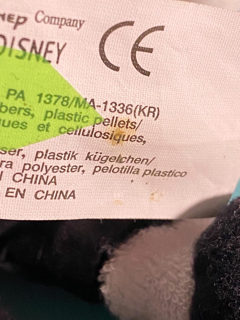 May include: Close-up of a white tag with the Disney logo and a CE mark. The tag includes text in multiple languages, including English, French, and Spanish, with the text "PA 1378/MA-1336(KR)" and "plastic pellets".