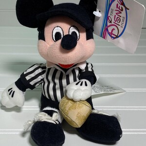May include: A plush Mickey Mouse doll wearing a black cap and a black and white striped shirt. The doll has white gloves and black shoes. It holds a heart-shaped object. A tag from The Disney Store is attached.