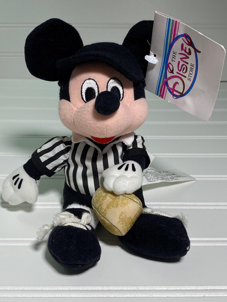 May include: A plush Mickey Mouse doll wearing a black cap and a black and white striped shirt. The doll has white gloves and black shoes. It holds a heart-shaped object. A tag from The Disney Store is attached.