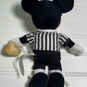 May include: A plush Mickey Mouse doll wearing a black and white striped referee shirt, black shorts, and black shoes. The doll has large black ears and white gloves. A tag is attached to the doll.