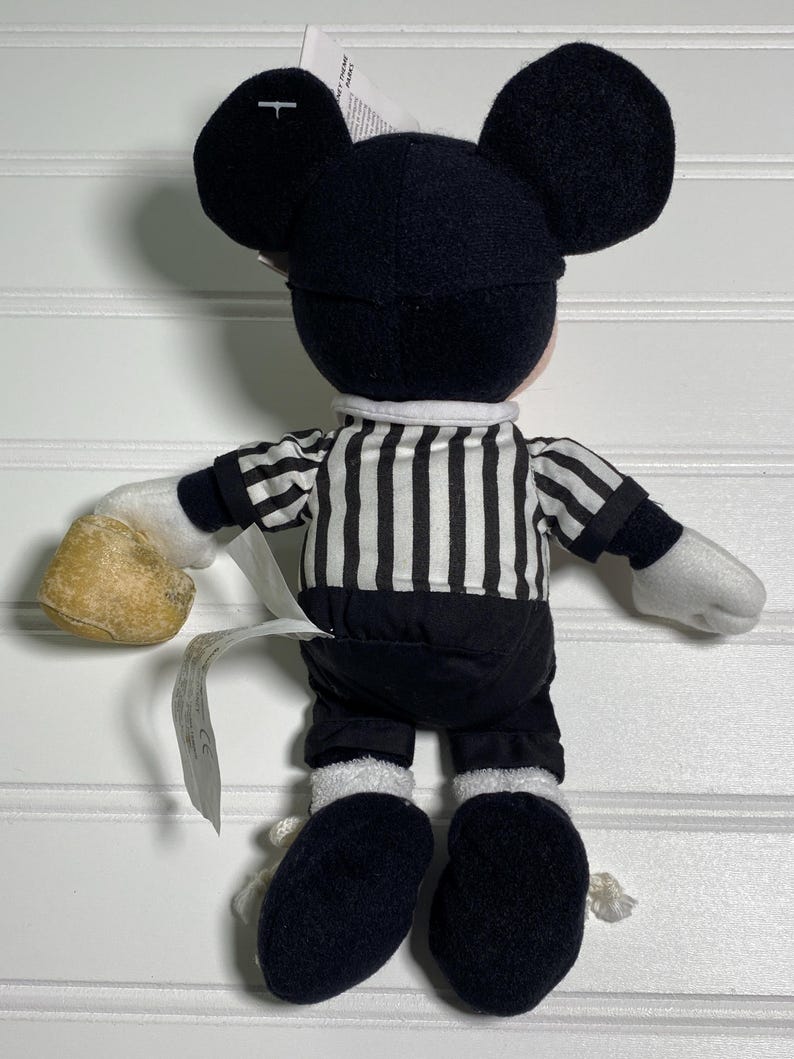 May include: A plush Mickey Mouse doll wearing a black and white striped referee shirt, black shorts, and black shoes. The doll has large black ears and white gloves. A tag is attached to the doll.