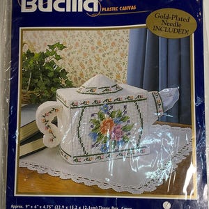 May include: A Bucilla plastic canvas kit to create a teapot tissue box cover. The white teapot-shaped box features a floral design in shades of blue, pink, and green. The kit includes a gold-plated needle and measures approximately 9" x 6" x 4.75".