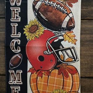May include: A decorative fall-themed garden flag with a football, red helmet, and pumpkin. The flag has the word "WELCOME" in white letters on a navy blue background. Sunflowers and autumn leaves complete the design.