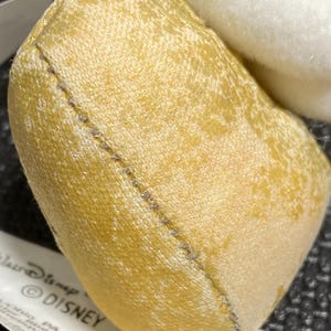 May include: Close-up of a small, heart-shaped plush toy in shades of yellow and cream. The toy has a stitched seam and a tag with the Disney logo. The plush is made of soft, textured fabric.