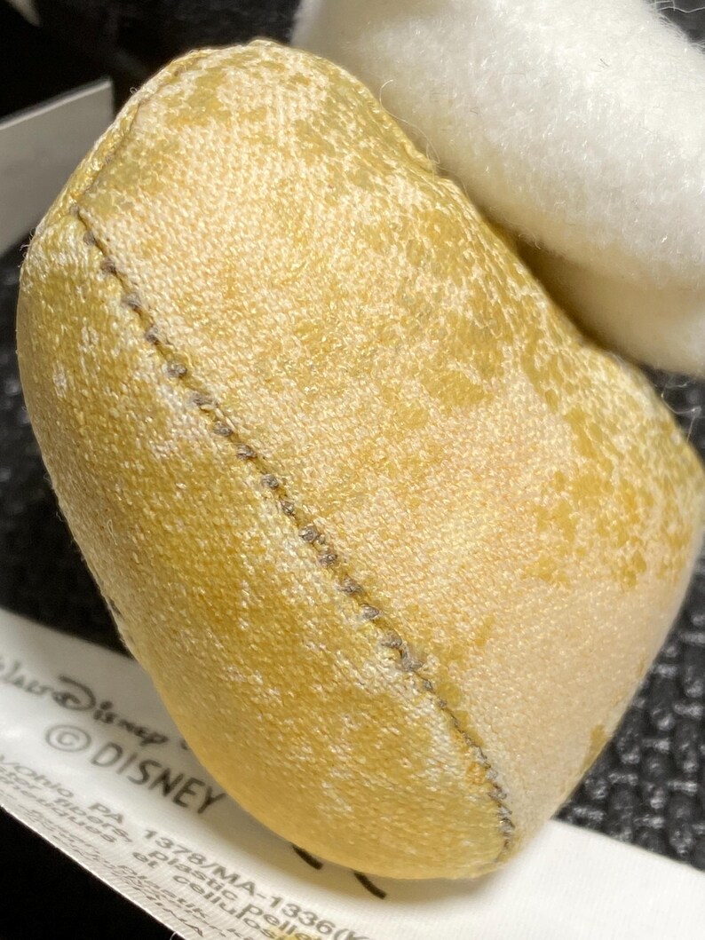 May include: Close-up of a small, heart-shaped plush toy in shades of yellow and cream. The toy has a stitched seam and a tag with the Disney logo. The plush is made of soft, textured fabric.