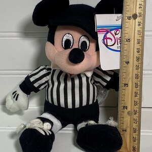 May include: A plush Mickey Mouse toy dressed as a referee, wearing a black and white striped shirt and shorts. The toy has a black hat and shoes, with a Disney Store tag attached. The toy measures approximately 7 inches tall.