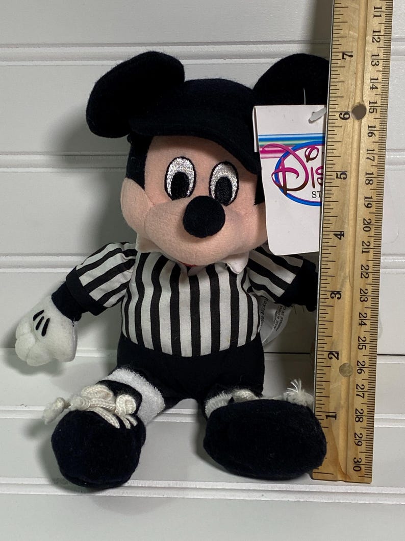 May include: A plush Mickey Mouse toy dressed as a referee, wearing a black and white striped shirt and shorts. The toy has a black hat and shoes, with a Disney Store tag attached. The toy measures approximately 7 inches tall.