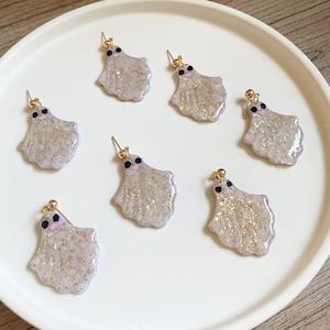 May include: Six pairs of gold dangle earrings with a white ghost design. Each ghost has black eyes and a pink blush. The ghosts are covered in glitter.
