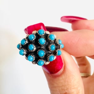 May include: A silver-toned ring featuring a cluster of small, round, bright turquoise stones. The ring has a detailed, textured design around each stone, creating a unique and eye-catching piece of jewelry. The ring is held up by a hand with red nail polish.