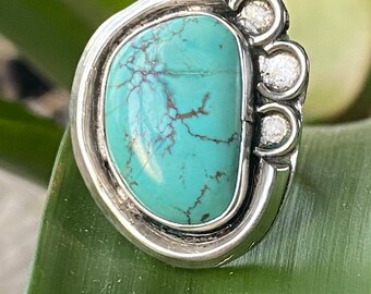 May include: A silver ring featuring a large, oval turquoise stone with brown veining. The ring has a unique design with three small, round accents on one side. The turquoise stone is a vibrant shade of blue-green.