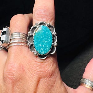 May include: A large, oval turquoise gemstone ring with a silver setting. The turquoise stone is a vibrant blue-green color, and the silver setting has a decorative, scalloped edge. Other rings are visible on the fingers.
