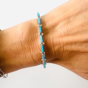 May include: A silver bracelet featuring a series of small, elongated, bright blue turquoise stones set vertically. Each stone is separated by a small silver bead, creating a delicate, repeating pattern along the length of the metal band.