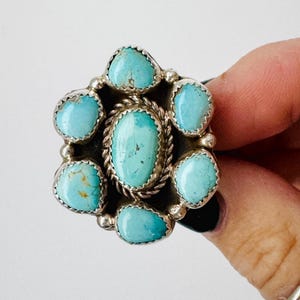 May include: A silver-toned ring featuring a cluster of turquoise stones. The ring has a central oval turquoise stone surrounded by six smaller, teardrop-shaped stones. The stones are set in a detailed, ornate silver setting, creating a floral-like design.