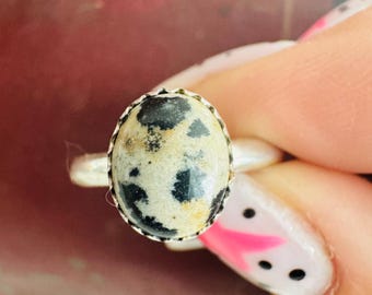 以下が含まれることがあります： 黒とクリーム色の斑点模様の石をあしらった楕円形のリング。シルバーカラーの金属バンドにセットされています。ピンクと白のマニキュアを塗った指でリングを持っています。石の長さは約2cmです。