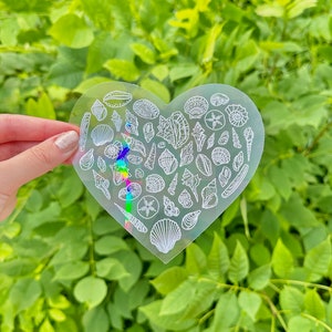 May include: A clear heart-shaped sticker with a holographic finish features a design of various seashells and starfish.