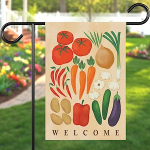 May include: A beige garden flag featuring a colorful illustration of various vegetables, including tomatoes, carrots, and peppers. The word "WELCOME" is printed in a bold, black font at the bottom of the flag. The flag is hanging on a black metal stand.