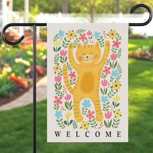 May include: A garden flag featuring a cheerful orange cat with its arms raised, surrounded by colorful flowers. The word "WELCOME" is printed in black at the bottom. The flag is hanging on a black metal stand in a garden setting.