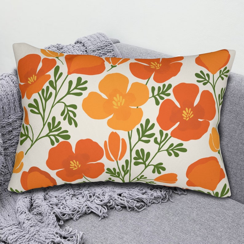 Pu&ograve; includere: Cuscino decorativo rettangolare con un motivo floreale di fiori arancioni e gialli con steli e foglie verdi su sfondo crema. Il cuscino &egrave; posto su un divano grigio con una coperta grigia.