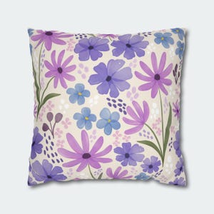 Può includere: Un cuscino quadrato con un motivo floreale. Il cuscino presenta uno sfondo crema con fiori viola, rosa e blu.