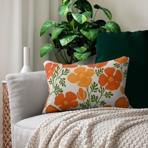 Pu&ograve; includere: Cuscino decorativo rettangolare con un motivo floreale di fiori arancioni e gialli con steli e foglie verdi. Il cuscino &egrave; su un divano grigio con una coperta color crema e un cuscino verde scuro.