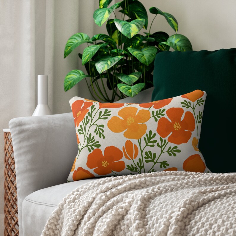 Pu&ograve; includere: Cuscino decorativo rettangolare con un motivo floreale di fiori arancioni e gialli con steli e foglie verdi. Il cuscino &egrave; su un divano grigio con una coperta color crema e un cuscino verde scuro.