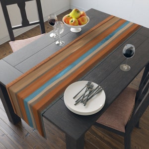 May include: A brown, beige, and blue striped table runner with a modern abstract design. The runner is laid on a dark wood table with a plate, silverware, and two wine glasses.