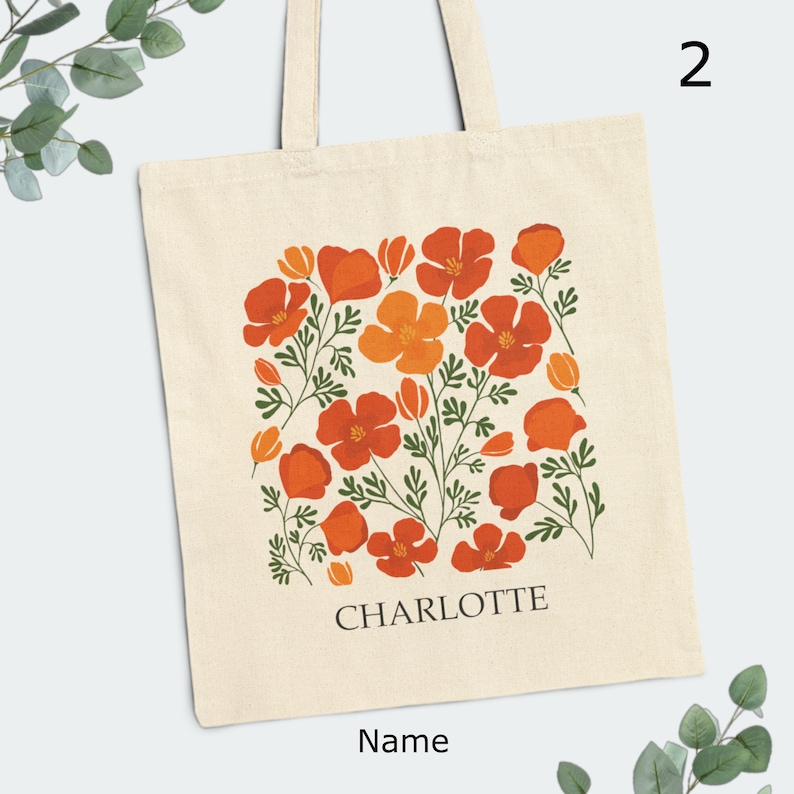 May include: A white canvas tote bag with a floral design featuring orange and red poppy flowers. The bag has a black text that says "CHARLOTTE" and "Name" below the design.