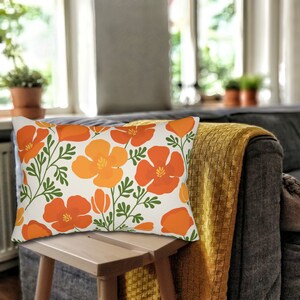 Pu&ograve; includere: Cuscino rettangolare decorativo con un motivo floreale di papaveri arancioni e gialli con foglie verdi su sfondo crema. Il cuscino &egrave; posto su uno sgabello di legno davanti a un divano grigio con una coperta gialla.