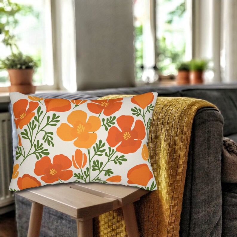 Pu&ograve; includere: Cuscino rettangolare decorativo con un motivo floreale di papaveri arancioni e gialli con foglie verdi su sfondo crema. Il cuscino &egrave; posto su uno sgabello di legno davanti a un divano grigio con una coperta gialla.