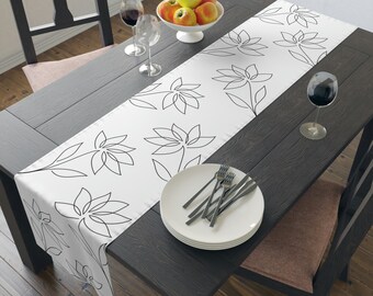 May include: White table runner with a black floral design. The runner is on a dark wood table with a bowl of apples, two wine glasses, and a plate with silverware.