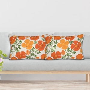 Pu&ograve; includere: Due cuscini rettangolari con un motivo floreale. Il design presenta fiori arancioni e gialli con steli e foglie verdi su sfondo crema. I cuscini sono posizionati su un divano grigio chiaro.