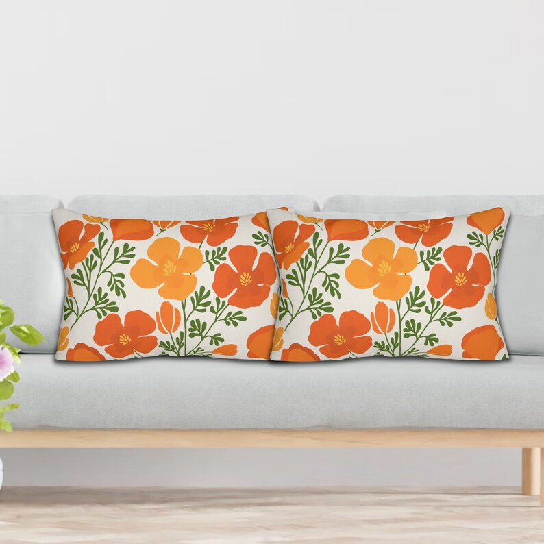 Pu&ograve; includere: Due cuscini rettangolari con un motivo floreale. Il design presenta fiori arancioni e gialli con steli e foglie verdi su sfondo crema. I cuscini sono posizionati su un divano grigio chiaro.