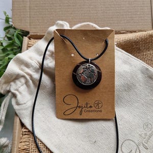 May include: A round pendant necklace featuring a world map design. The pendant hangs on a black cord and is displayed on a brown card with the "Jojito Creations" logo. The necklace is presented on a white cloth bag.