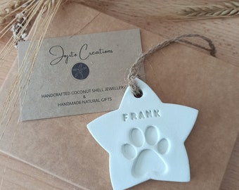May include: A white star-shaped ornament featuring an embossed paw print and the name FRANK, suspended by a brown twine loop. The ornament is placed next to a brown card with the text Jojito Creations, HANDCRAFTED COCONUT SHELL JEWELLERY & HANDMADE NATURAL GIFTS.