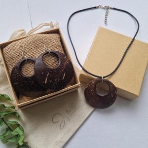 May include: A jewellery set comprising a necklace and earrings made from dark brown coconut shells. The earrings are circular with a hole in the centre, displayed in a box. The necklace has a matching pendant on a black cord. A small beige bag is also present.