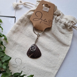 May include: A brown, triangular keychain pendant with a silver wave design, attached to a silver chain and keyring. The pendant is made of a dark, natural material. The tag reads "Jojita Creations". The keychain rests on a beige drawstring bag.