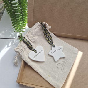 May include: Two white ceramic ornaments, one shaped like a heart and the other a star, both with the word "Cornwall" written on them. The ornaments are in a small white drawstring bag with a yellow and black checked ribbon.