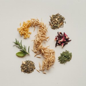 May include: An overhead shot displays various dried herbs and grains arranged on a white surface. The assortment includes golden-yellow flower buds, brown grains, green rosemary, and dark red hibiscus petals. The arrangement suggests ingredients for teas or culinary use.