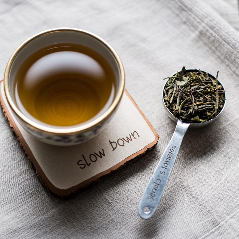 May include: A teacup with a gold rim on a wooden coaster with the words "slow down". A metal measuring spoon filled with loose tea leaves is next to the cup. The tea is a light amber colour.